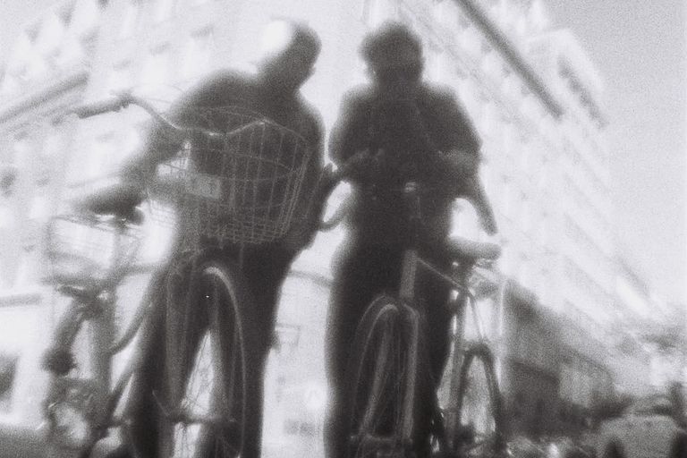 Blurry black and white photo of electronic music duo Poltergeist 9000 posing with bicycles outdoors against a washed-out city building background.